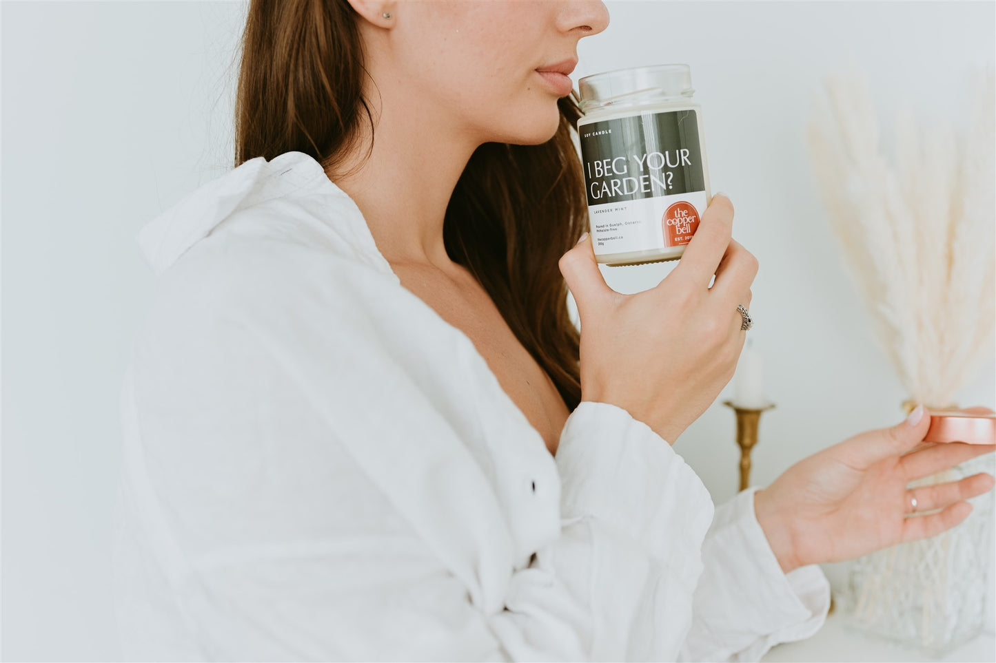 woman wearing white shirt smelling i beg your garden candle. smells like a relaxing blend of lavender and mint.